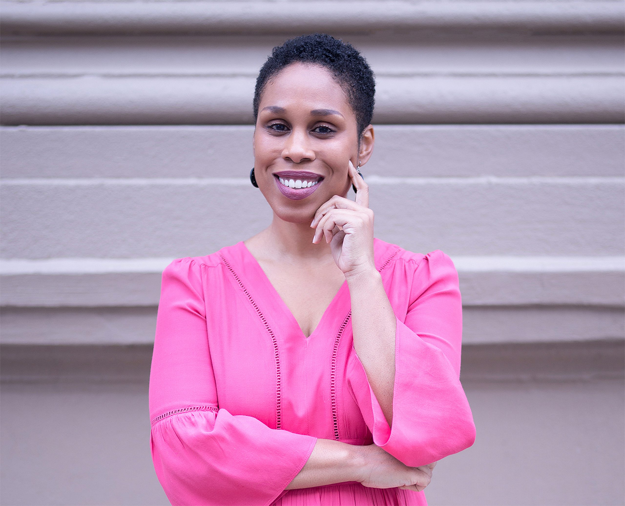 Dr. Oni Blackstock standing with her arms folded, her index finger pressing into her cheek, wearing a bright pink blouse