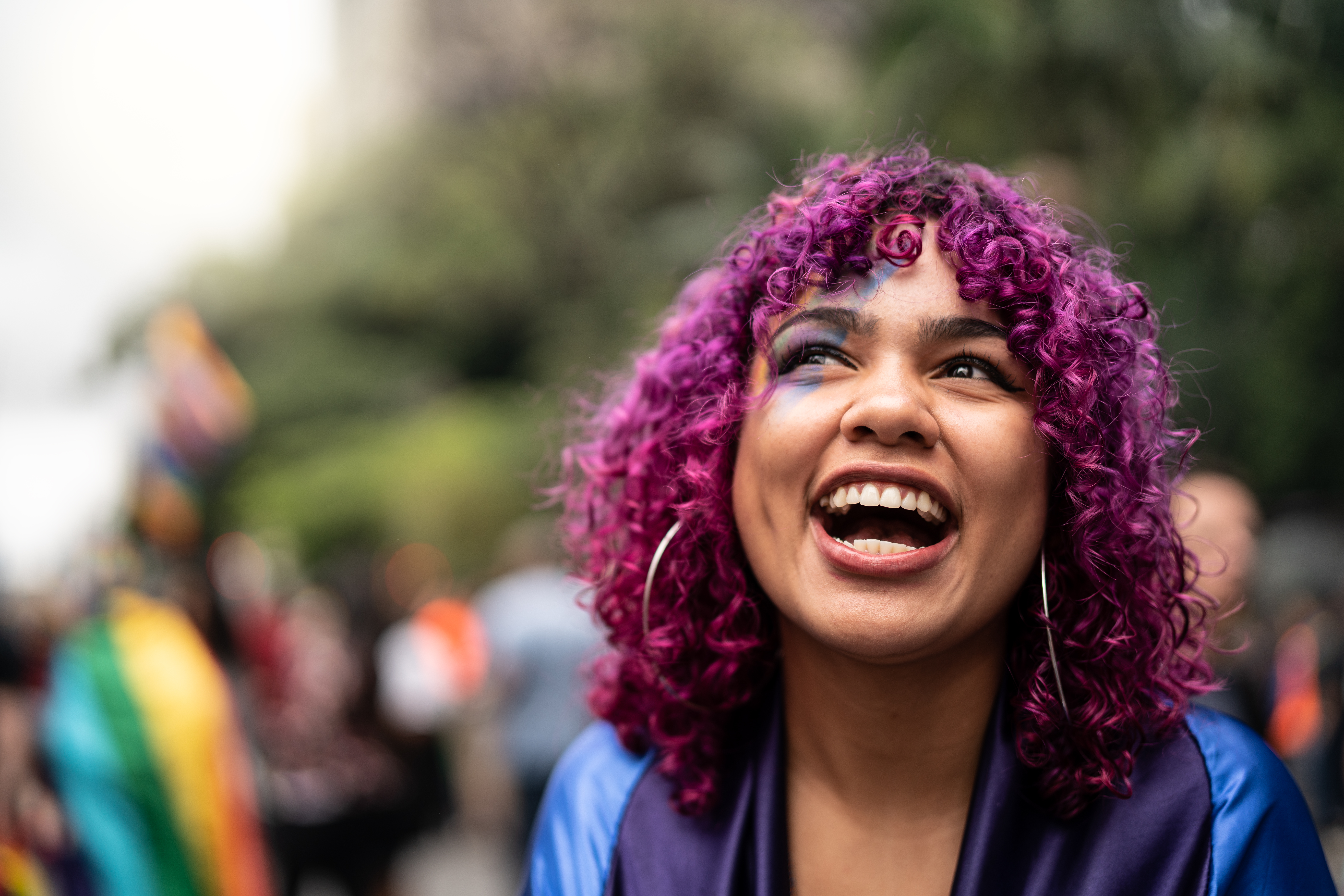 A young woman of color with curly purple hair and a painted face smiles at pride parade